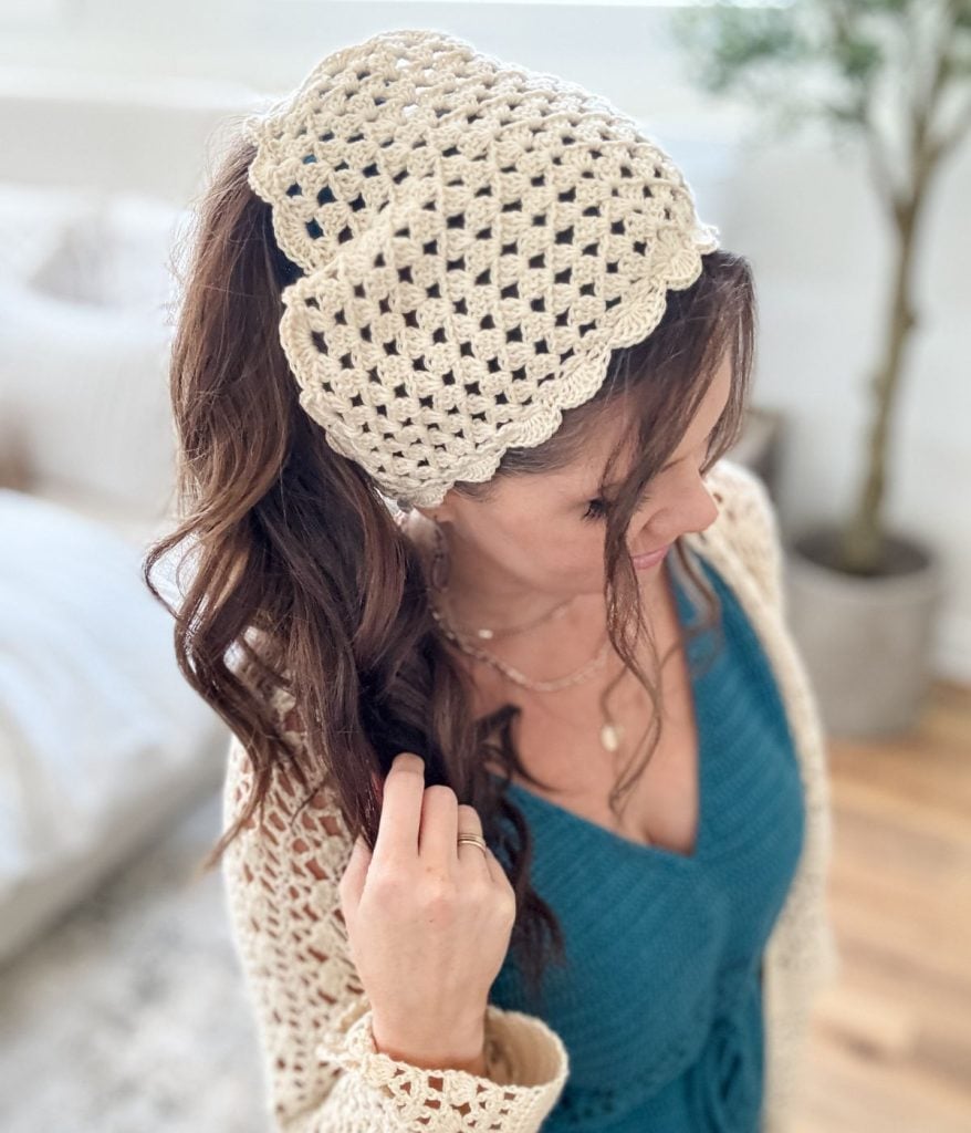 Woman with long brown hair wears a stylish crochet bandana as a cream headscarf and a blue top, standing indoors with neutral décor in the background.