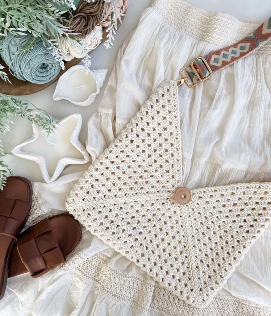 A cream Caribbean Beach Bag with a button closure rests on a white skirt, next to brown sandals, candles, and skeins of yarn. Greenery decorates the scene.