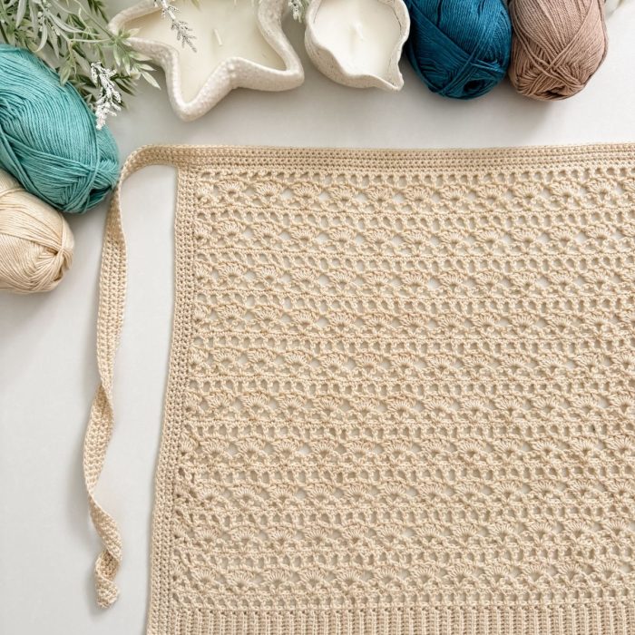 Rectangular crocheted lace placemat, inspired by a Caribbean Seashell crochet pattern, with a tie on one side, surrounded by balls of yarn, candles, and white foliage on a white surface.