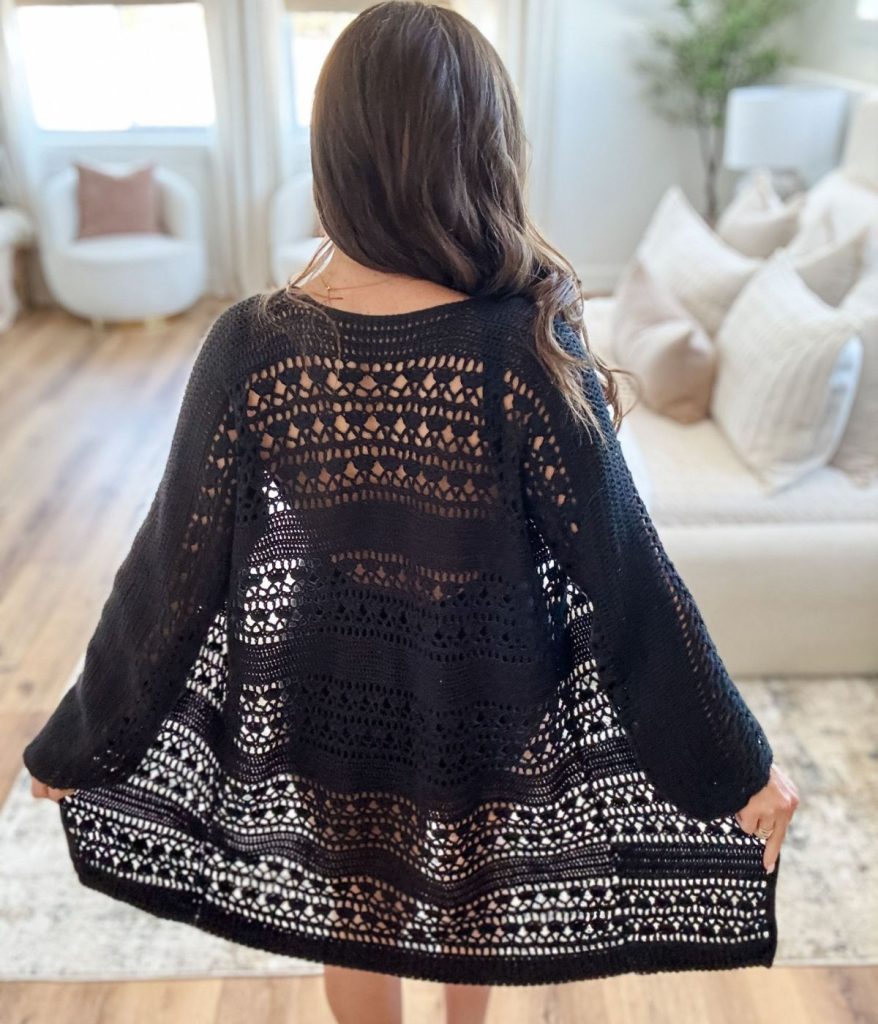 A woman with long brown hair stands indoors facing away, wearing a black Caribbean Crochet Cardigan spread out to display its intricate open-knit pattern.