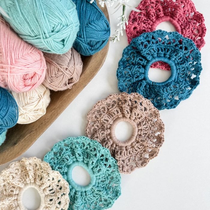 Five crocheted hair scrunchies in various colors are displayed in a row next to a wooden bowl filled with yarn skeins in matching shades, perfect for pairing with your favorite Caribbean Crochet Cardigan.