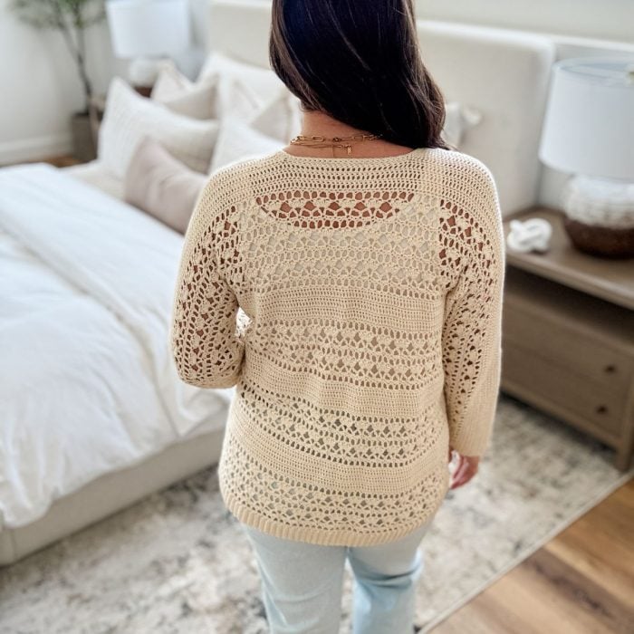 A woman with long brown hair, seen from behind, wears a beige Caribbean Crochet Cardigan and light jeans as she stands next to a neatly made bed in a bright, modern bedroom.