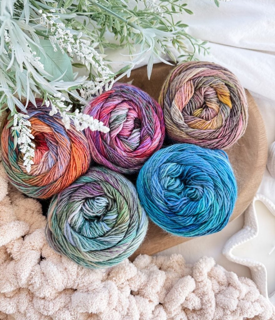 Five multicolored yarn balls arranged on a wooden tray surrounded by green foliage and cream-colored textured fabric—perfect inspiration for your next Caribbean Crochet Cardigan project.