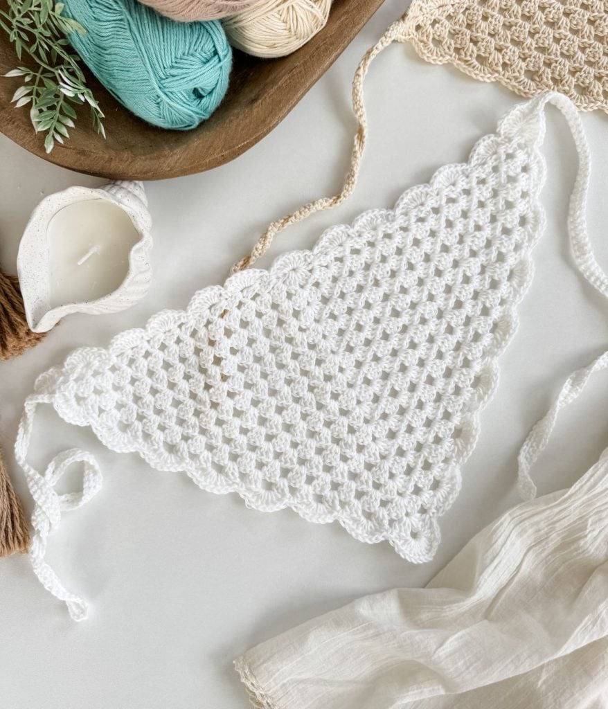 A Crochet Bandana in white, featuring a scalloped edge, is displayed on a table surrounded by yarn, a crochet piece, a candle, and fabric.