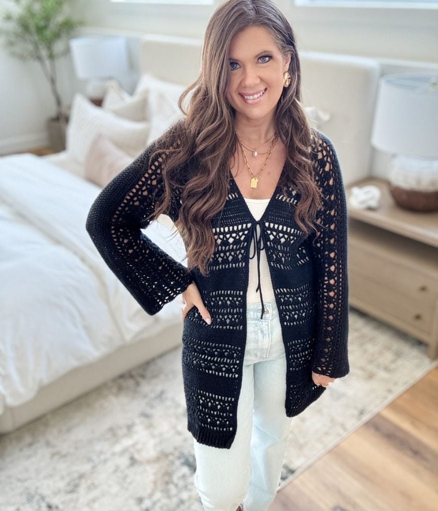 Woman standing in a bedroom, smiling at the camera, wearing a white top and light jeans topped with a stylish black Caribbean Crochet Cardigan.