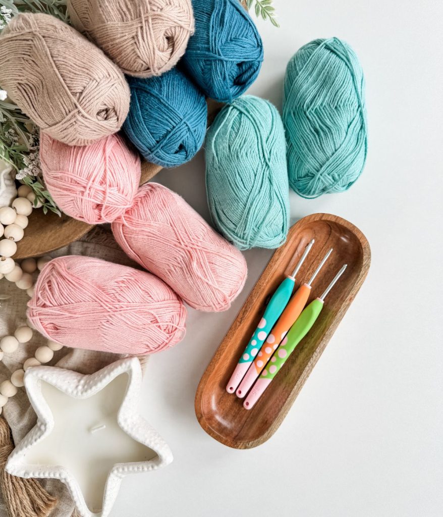 Several skeins of pastel yarn, three crochet hooks in a wooden tray, a white star-shaped candle, and wooden beads rest on a white surface—perfect essentials for crafting your own Caribbean Crochet Cardigan.