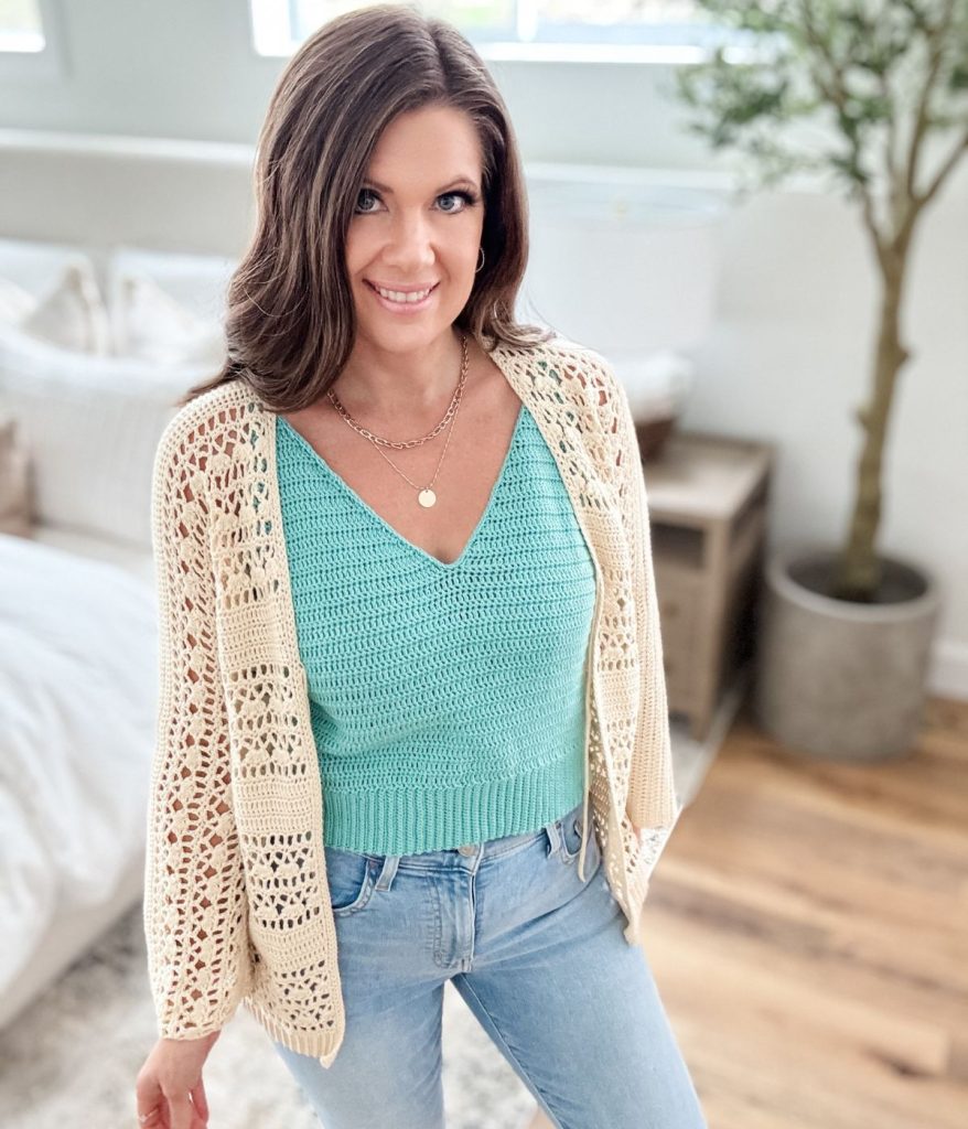 A woman with long brown hair is wearing a turquoise crochet tank top, a cream cardigan, and light blue jeans, standing in a bright, modern bedroom.