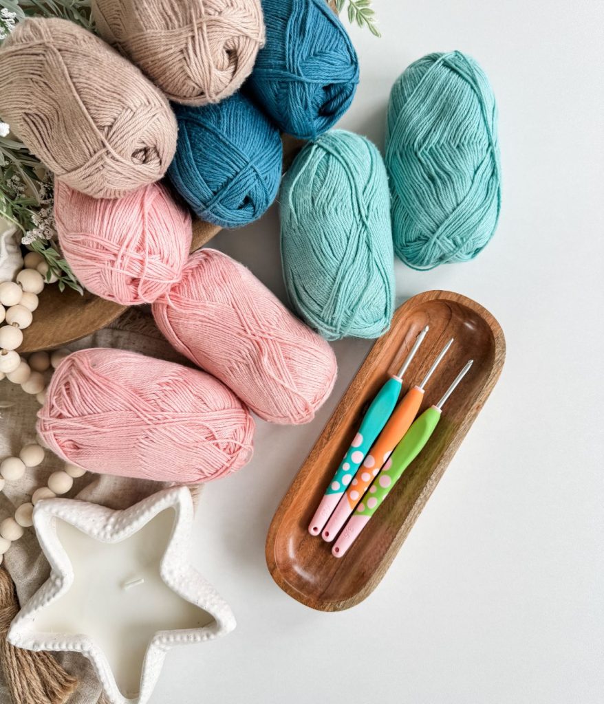 Several skeins of yarn in pink, teal, blue, and beige are arranged with four crochet hooks in a wooden tray, alongside a white star-shaped candle—perfect inspiration for crafting your own Caribbean Crochet Cardigan.
