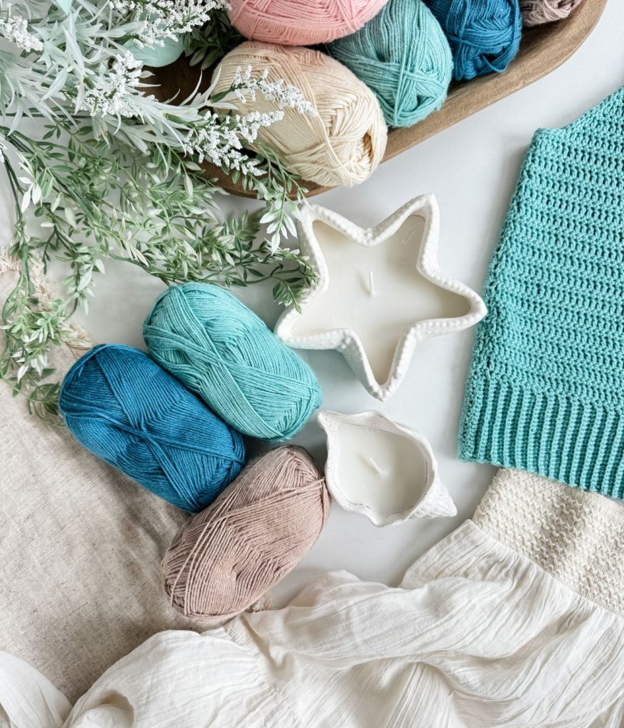 Yarn balls, a knitted fabric, two white candles in ceramic holders, and green foliage are arranged on a white surface with beige cloth—perfect inspiration for your next crochet tank top project.
