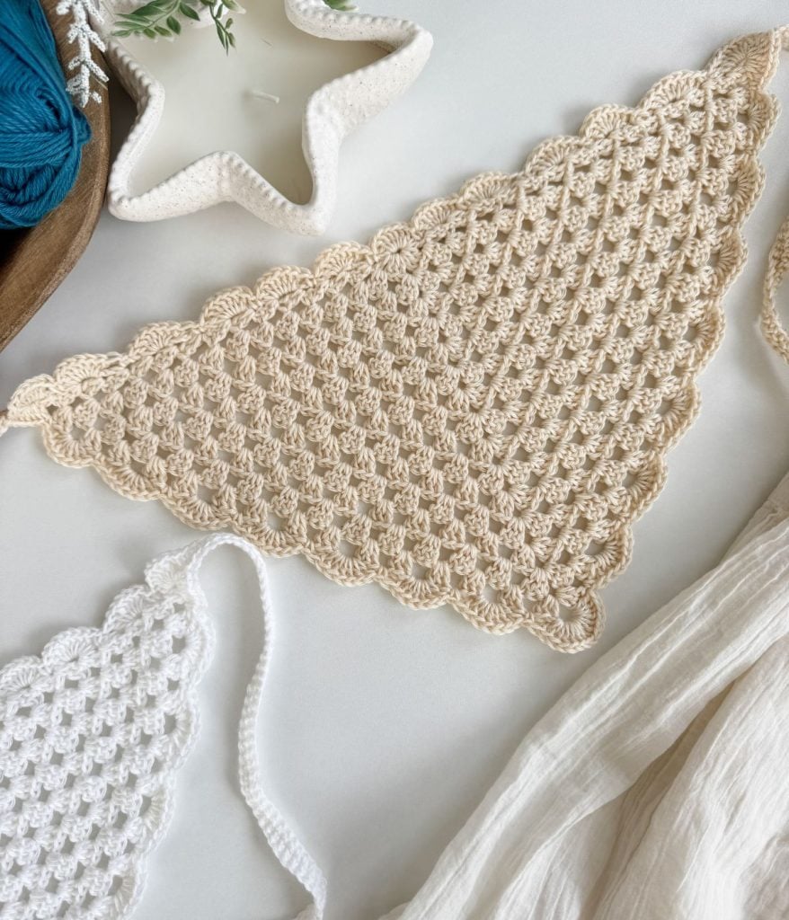 A beige crochet triangle bandana with a scalloped edge lies on a white surface next to a white crochet piece, a star-shaped dish, and a light fabric.