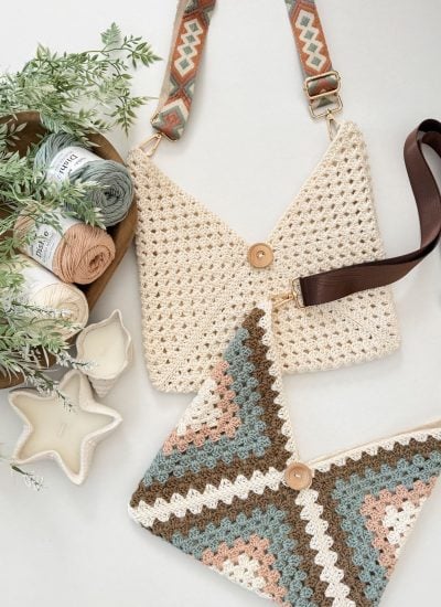 Two crochet crossbody bags with button closures lie on a surface, next to skeins of yarn, greenery, and two star-shaped candles—perfect as stylish beach bags for your summer outings.