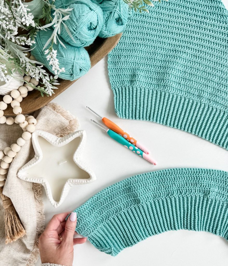 A hand holds a partially crocheted teal piece next to a finished crochet tank top, yarn balls, crochet hooks, a star-shaped candle, beads, and greenery arranged on a white surface.