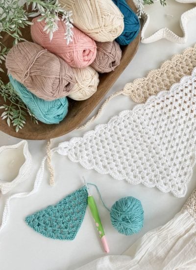 A collection of colorful yarn balls, a crochet hook, and triangular crochet pieces—perfect for making a stylish crochet bandana—are arranged on a white surface with decorative greenery.