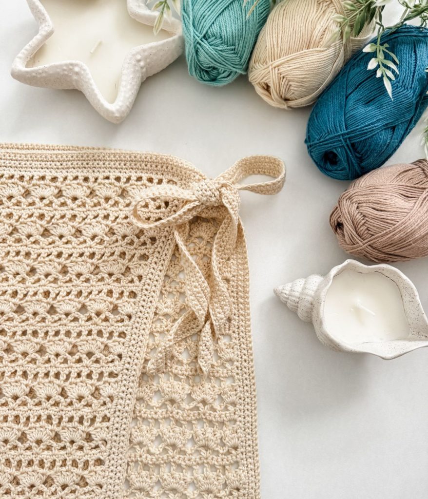 A crocheted beige fabric with a tied bow is shown beside yarn balls in various colors, two candles in decorative holders, and a Skirt Crochet Pattern inspired by Caribbean Seashell hues.