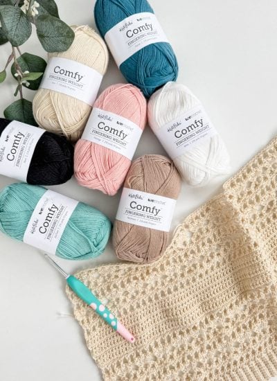 Seven skeins of cotton yarn, a teal crochet hook, and a partially crocheted cream-colored fabric—perfect for your next Caribbean Crochet Cardigan—are arranged on a white surface with green leaves in the corner.