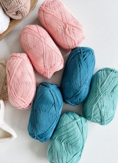 Seven skeins of yarn in shades of pink, blue, and teal are arranged on a white surface near a star-shaped dish and woven fabric—perfect hues for your next crochet tank top project.