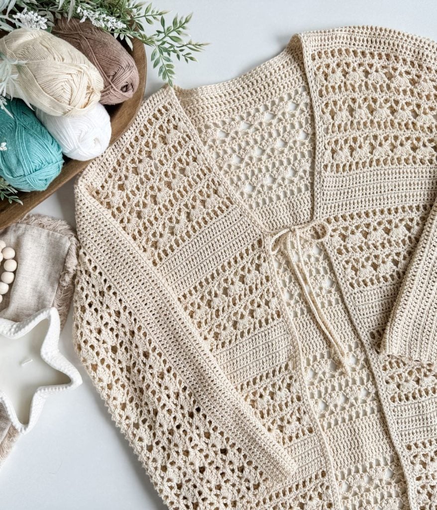 A beige Caribbean Crochet Cardigan is laid flat next to a wooden tray with balls of yarn in cream, brown, green, and white shades. A white star-shaped dish is also visible.