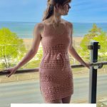 A woman wearing a pink crochet dress stands on a balcony overlooking a beach and green trees under a blue sky. Text reads: "Caribbean Cocktail Coverup Crochet Pattern & Granny Square Bag Video.