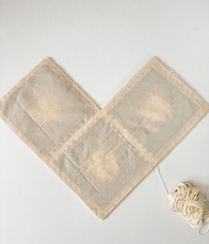 Two rectangular pieces of beige fabric are sewn together in an L-shape, resembling the base of a Granny Square Bag, with a ball of cream-colored yarn attached to one edge.