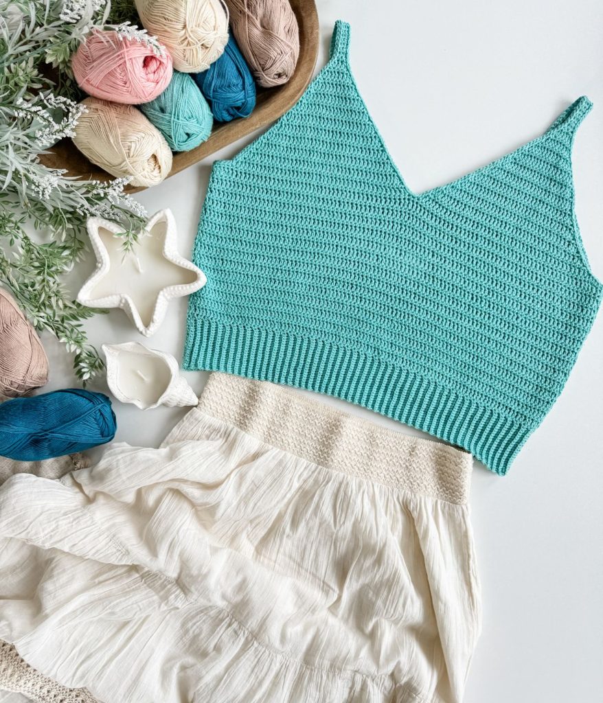 A crocheted turquoise crop top and cream skirt are laid flat beside yarn balls, a wooden tray, and ceramic star-shaped dishes on a white surface, showcasing the handmade charm of this Crochet Tank Top ensemble.