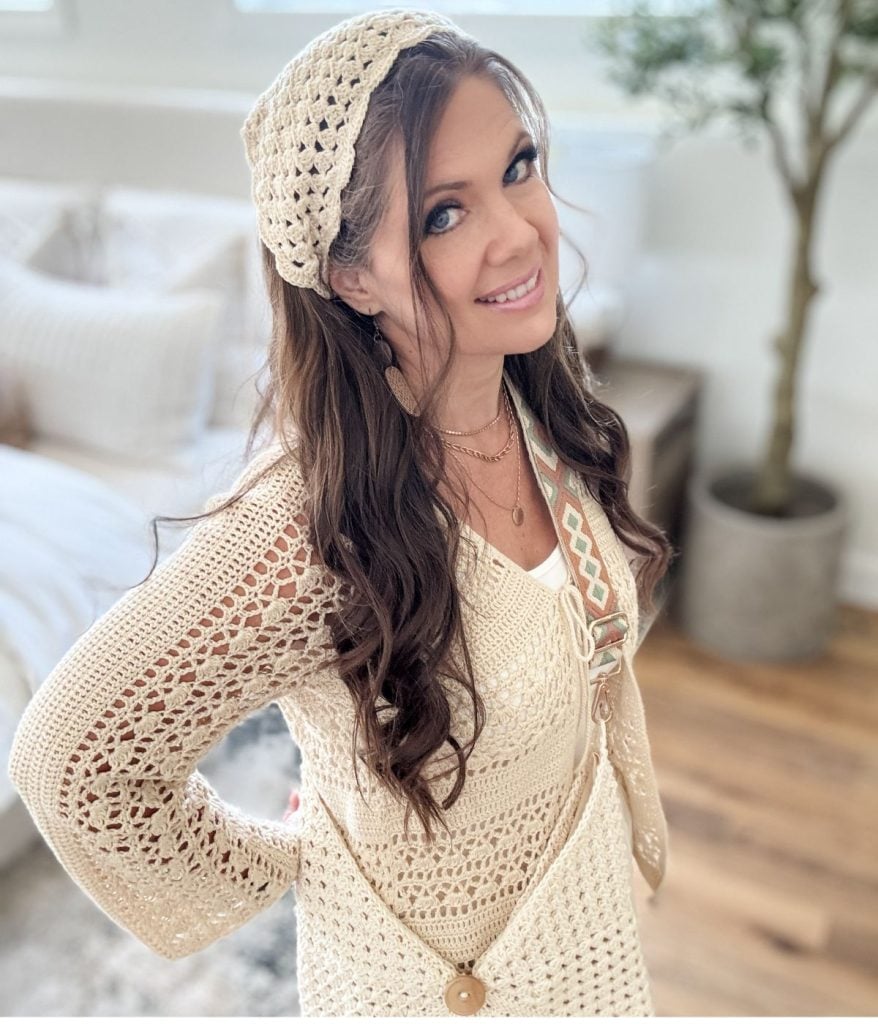 Woman with long brown hair wearing a cream-colored crocheted top, matching crochet bandana headband, and crossbody bag stands indoors, smiling at the camera.