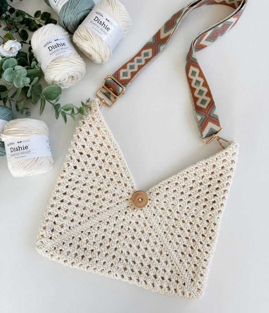 A cream-colored crocheted envelope bag inspired by Caribbean crochet, featuring a button closure and patterned strap, displayed next to four skeins of Dishie yarn and some greenery.