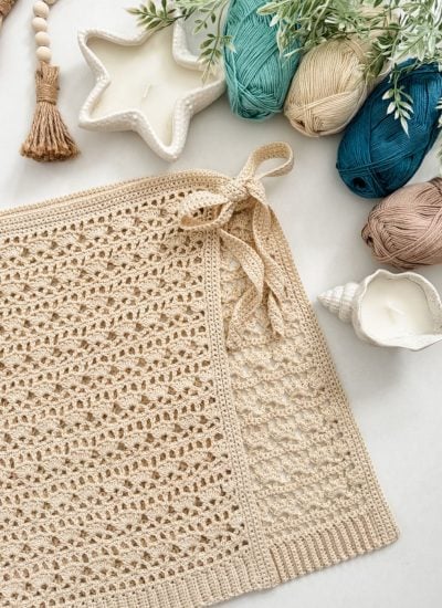 A beige crocheted rectangle with a bow, inspired by a Caribbean Seashell crochet pattern, lies next to yarn balls, star and shell-shaped candles, greenery, and decorative tassels on a light surface.