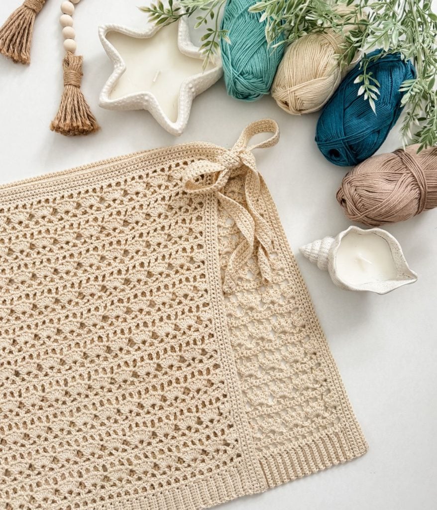A beige crocheted rectangle with a bow, inspired by a Caribbean Seashell crochet pattern, lies next to yarn balls, star and shell-shaped candles, greenery, and decorative tassels on a light surface.