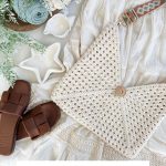 Flat lay of a white granny square bag with a wooden button, a white skirt, brown sandals, starfish, candles, and greenery on a light background. Text reads "Caribbean Beach Bag.