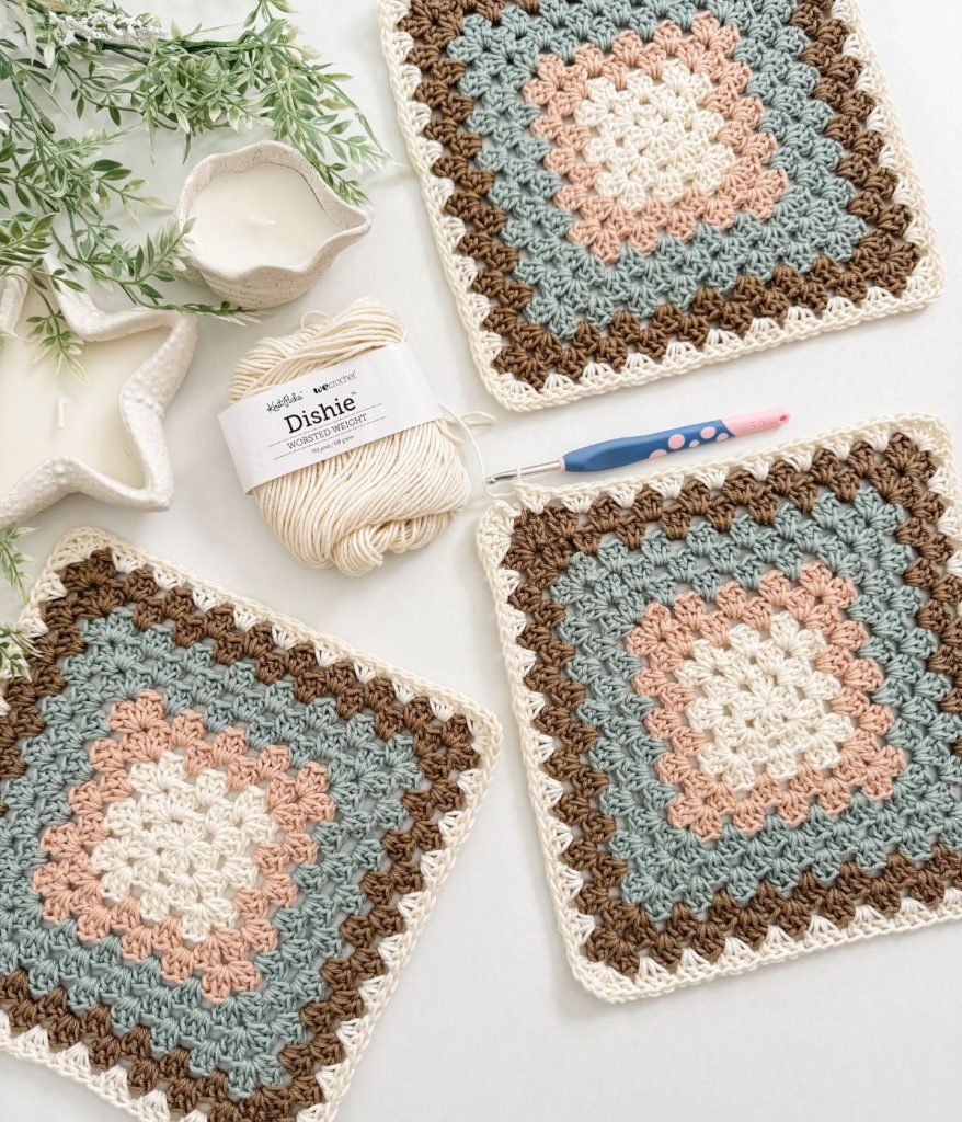 Three crocheted granny squares in pastel colors are displayed alongside a pink crochet hook, cream Dishie yarn, and a decorative plant—perfect inspiration for your next granny square bag or crochet pattern project.