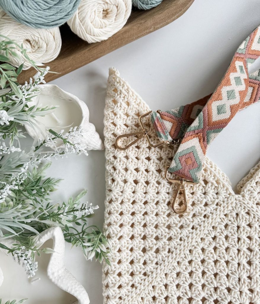A close-up of a crocheted cream Granny Square Bag with a patterned strap, surrounded by yarn balls, lace, and green foliage on a white surface.