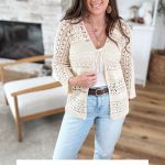 Woman wearing a cream-colored Caribbean Crochet Cardigan over a white top and light blue jeans, standing in a modern living room. Text reads: "CARIBBEAN BREEZE CARDIGAN Sizes XS-5X.