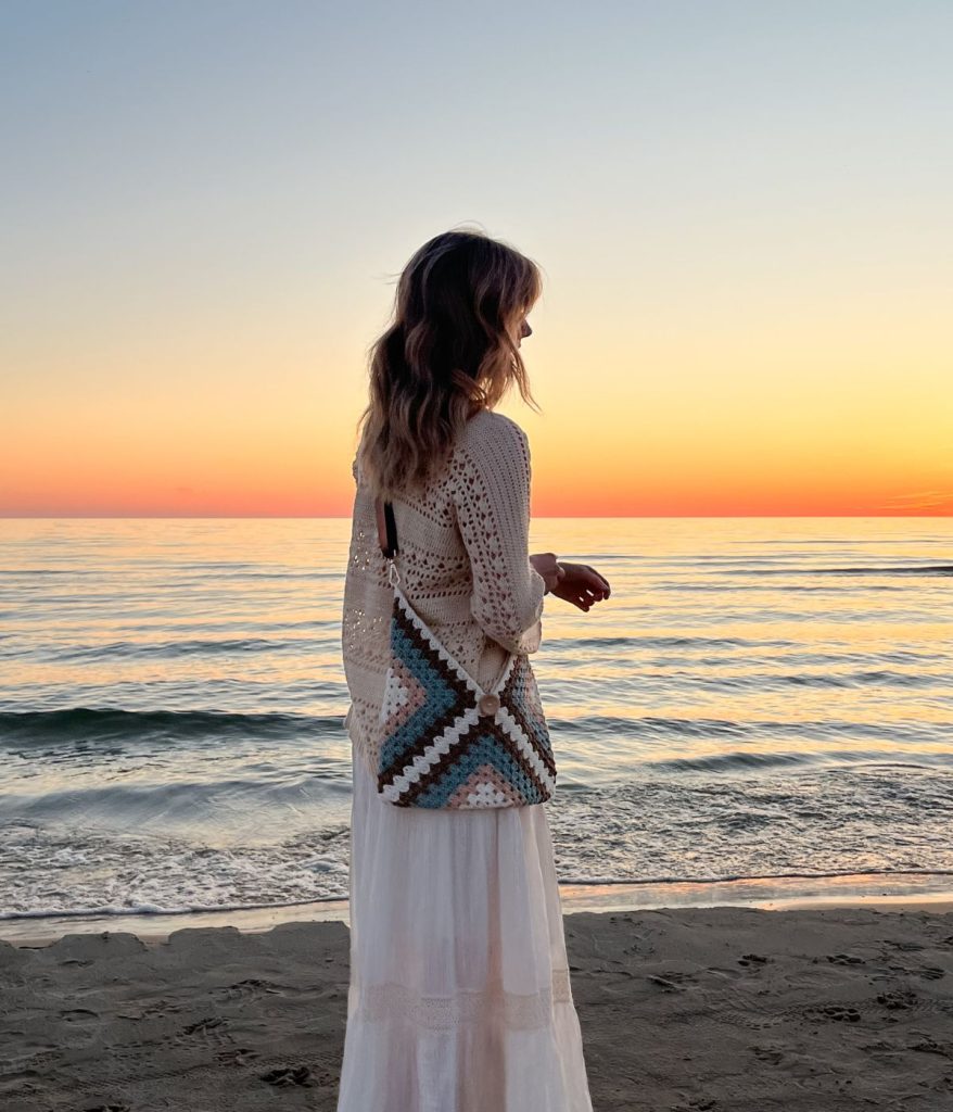 A woman in a white dress with a granny square bag stands on the beach at sunset, gazing out toward the ocean.