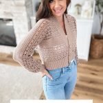 Woman wearing a light brown Caribbean Crochet Cardigan and light blue jeans, standing in a modern living room. Text reads: "Caribbean Crop Cardigan, Sizes XS-5X.