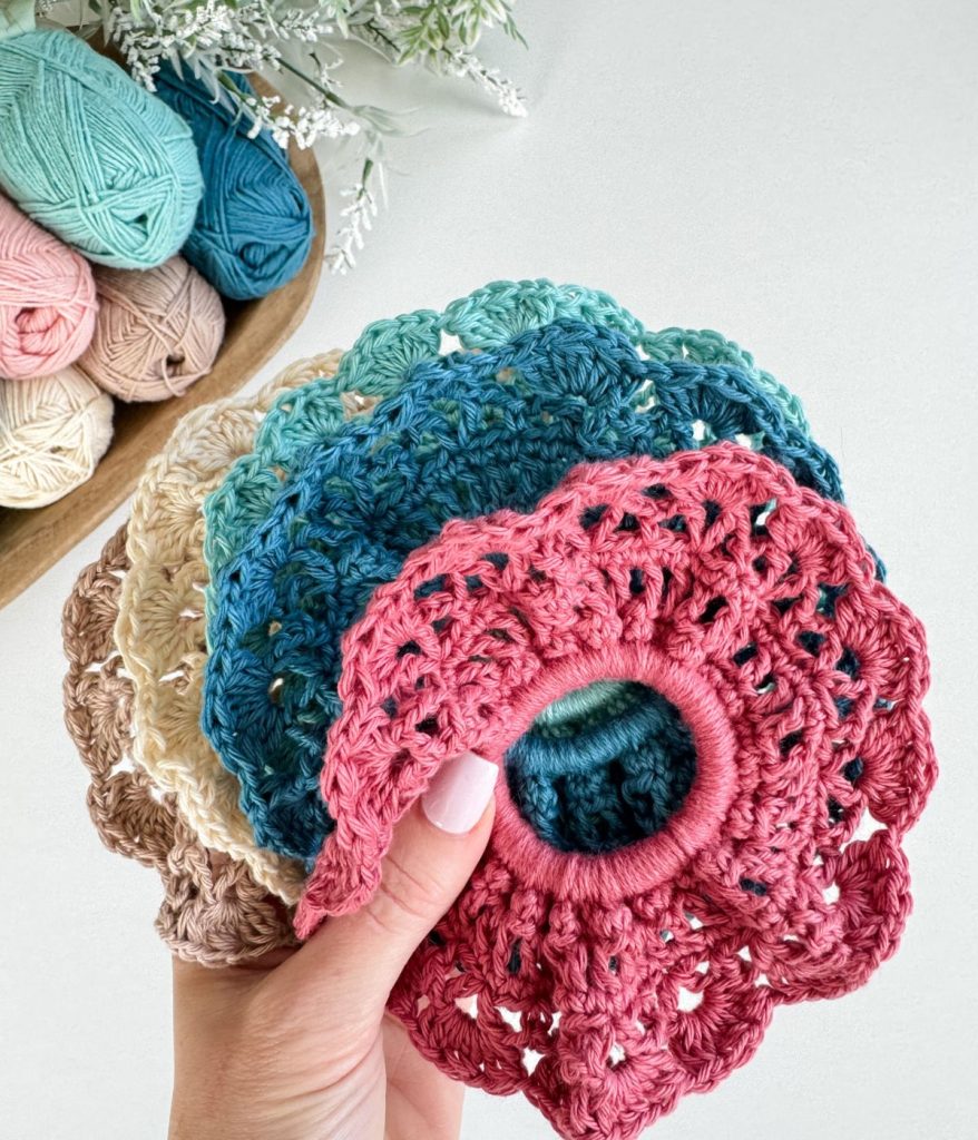 A hand holds several crocheted floral coasters in various colors, with yarn balls and greenery visible in the background, reminiscent of the vibrant hues found in a Caribbean Crochet Cardigan.
