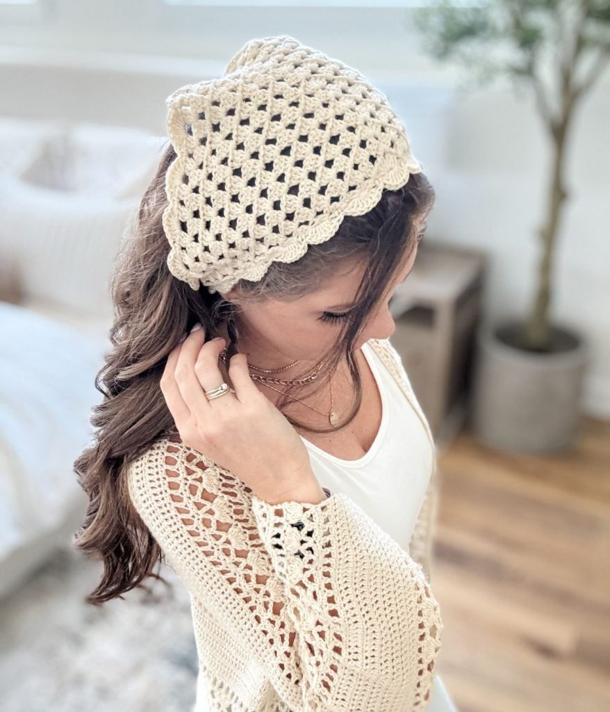 Woman with long brown hair wearing a white crochet bandana headscarf and matching crochet cardigan, standing indoors on a wooden floor.