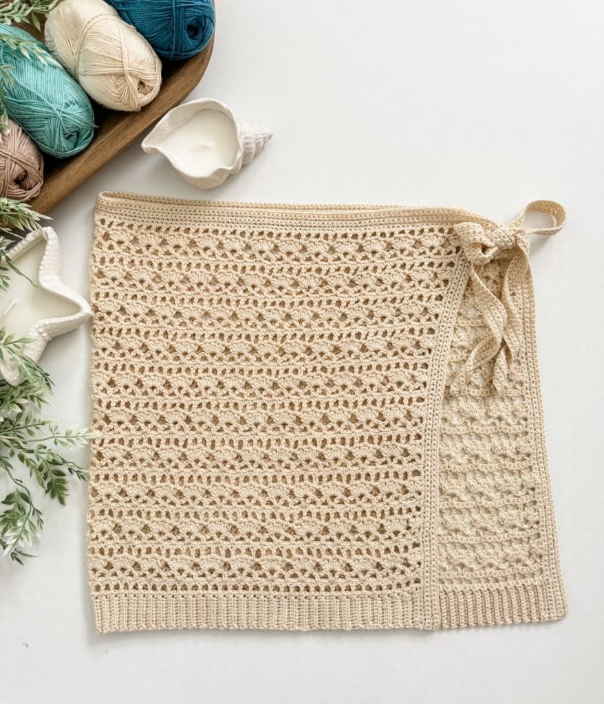 A crocheted beige wrap skirt, crafted with a Caribbean Seashell crochet pattern and a tie on one side, is displayed flat on a white surface beside yarn balls and greenery.