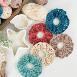 Five crocheted hair scrunchies in various colors are arranged on a table with yarn balls, shell-shaped candles, and decorative beads—perfect for pairing with your favorite Caribbean Crochet Cardigan.