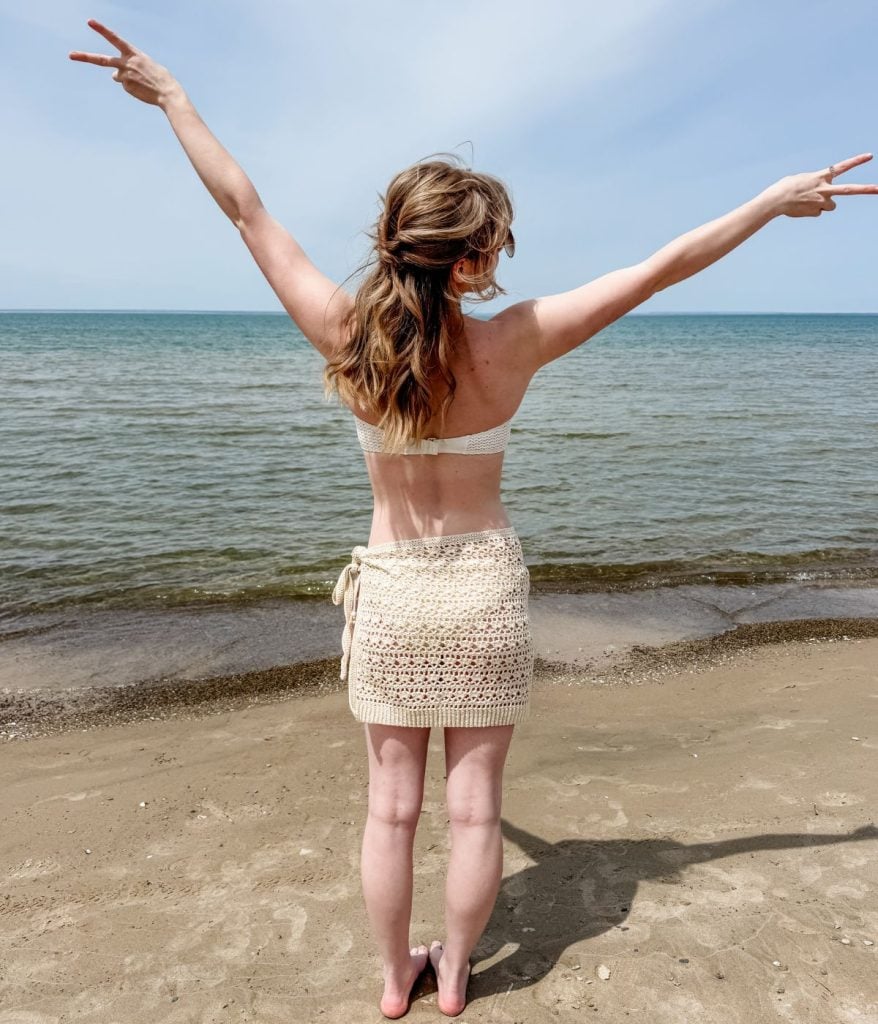 A woman stands barefoot on a sandy beach facing the water, arms raised with peace signs in both hands, her skirt crochet pattern flowing gently in the sea breeze.