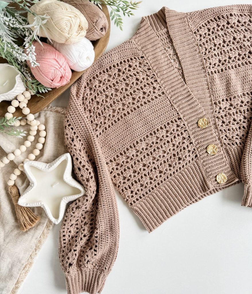 A light brown Caribbean Crochet Cardigan with gold buttons is displayed on a white surface next to balls of yarn, a beaded garland, a star-shaped dish, and a sprig of greenery.