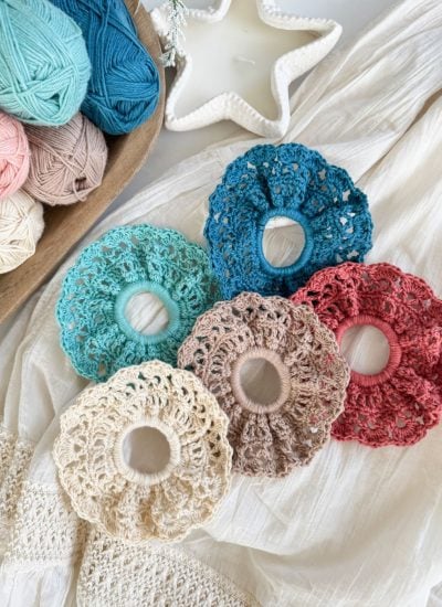 Five crocheted hair scrunchies in various colors are displayed on a cream fabric surface beside balls of yarn and a star-shaped ceramic dish, perfect for pairing with your favorite Caribbean Crochet Cardigan.