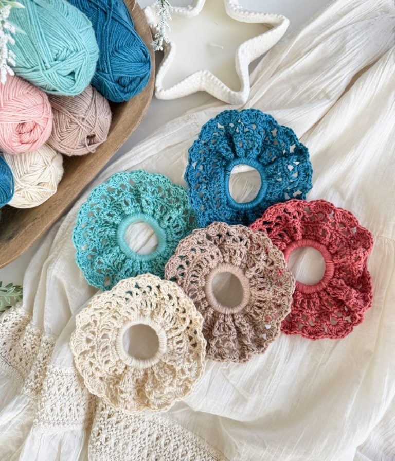 Five crocheted hair scrunchies in various colors are displayed on a cream fabric surface beside balls of yarn and a star-shaped ceramic dish, perfect for pairing with your favorite Caribbean Crochet Cardigan.