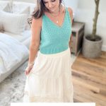 Woman wearing a light blue Crochet Tank Top and white skirt stands in a bright, cozy room; text reads "Caribbean Tide Tank Top Crochet Pattern & Video.