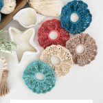 Five intricately made crochet scrunchie hair ties in various colors are arranged on a white surface, surrounded by yarn balls and a star-shaped ceramic dish.