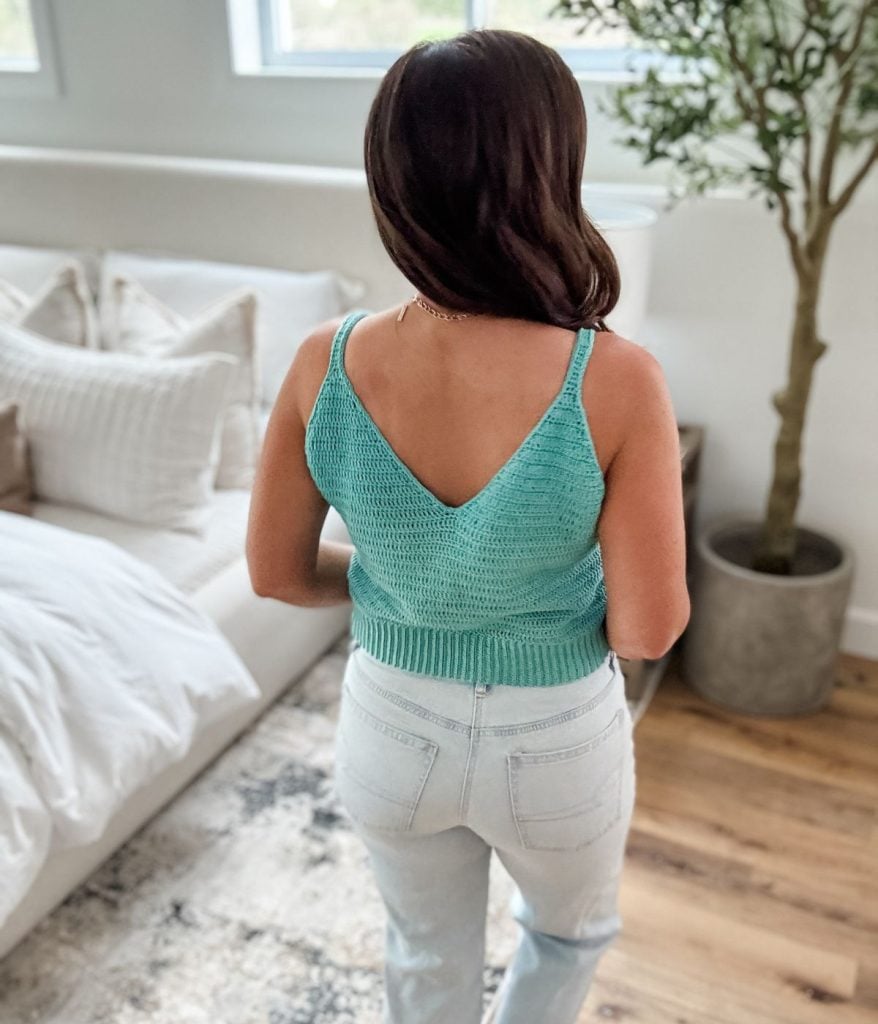 A woman with long brown hair stands indoors, facing away, wearing a teal crochet tank top and light blue jeans. The room features a bed, pillows, a cozy rug, and a potted plant.
