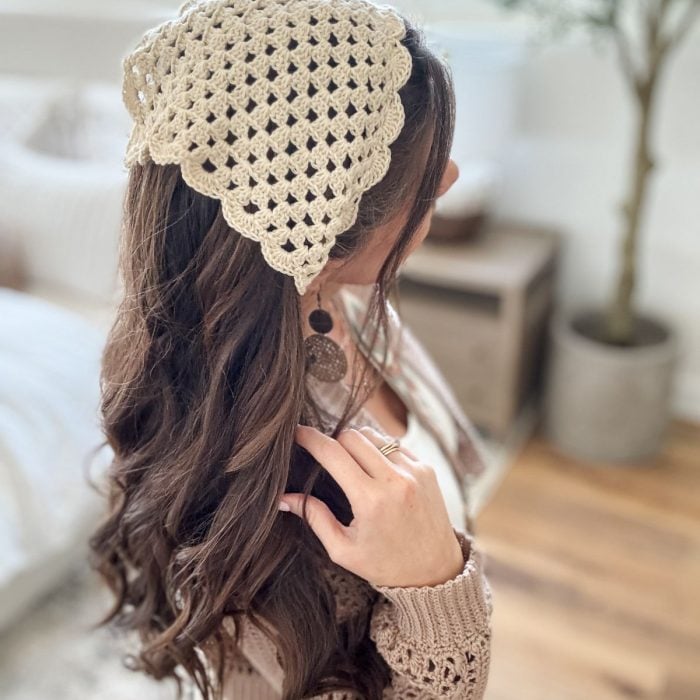 A woman with long brown hair wears a beige crochet bandana and a beige sweater, standing indoors near a potted plant and wooden furniture.
