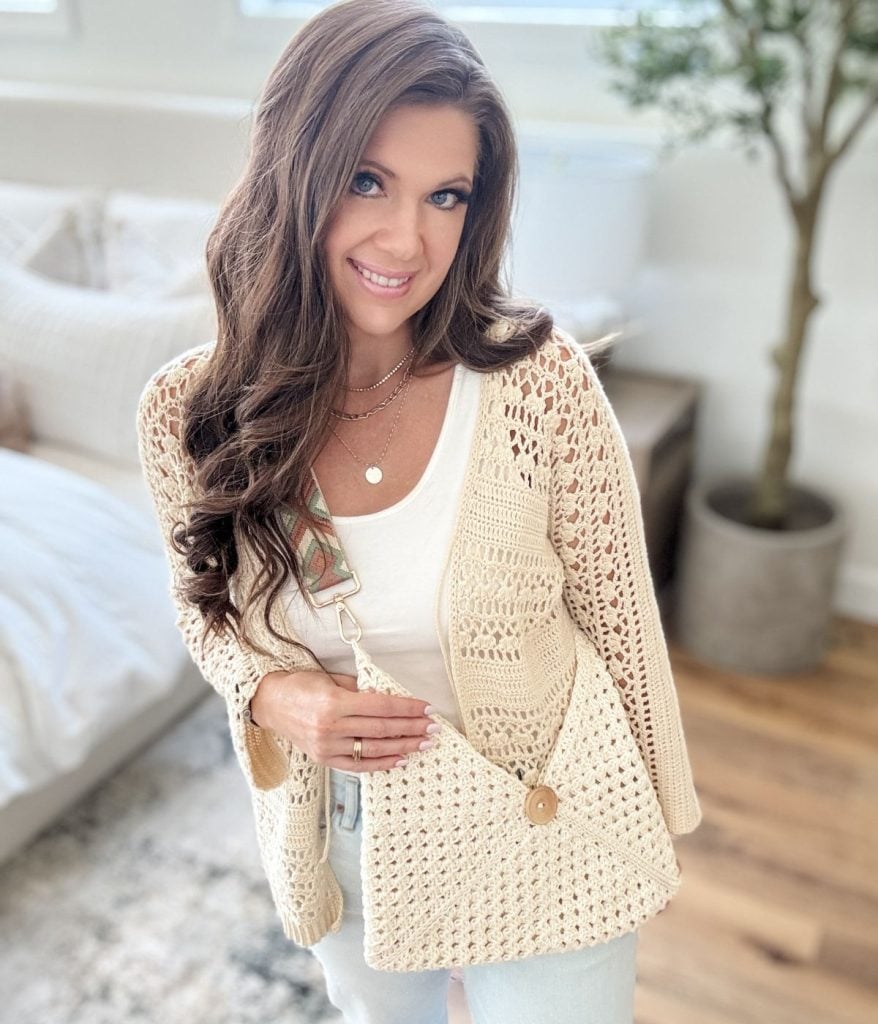 Woman with long brown hair wearing a cream crochet cardigan and a chic granny square bag, standing in a bright room with a plant in the background.