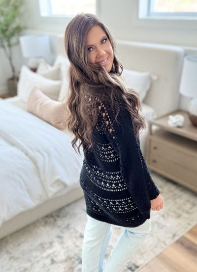 A woman with long brown hair wearing a Caribbean Crochet Cardigan and light jeans stands in a bright bedroom, looking over her shoulder and smiling.