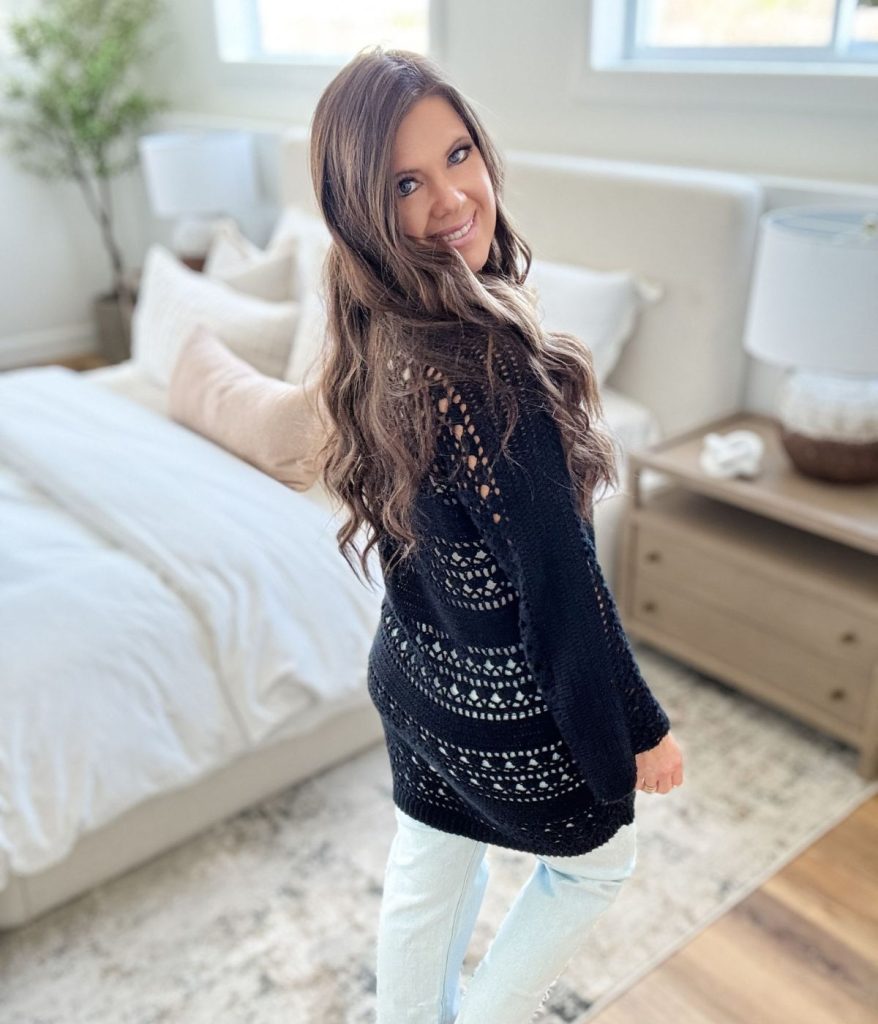 A woman with long brown hair wearing a Caribbean Crochet Cardigan and light jeans stands in a bright bedroom, looking over her shoulder and smiling.