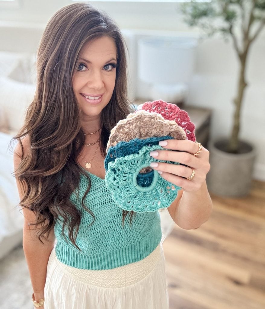 A woman with long brown hair holds up a set of colorful crocheted scrunchies in a bright, cozy room, her Caribbean Crochet Cardigan adding a vibrant touch to the scene.